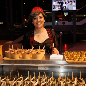 A smiling woman wearing a red headscarf and traditional attire stands behind a table filled with beautifully presented Middle Eastern dishes at a catering event.